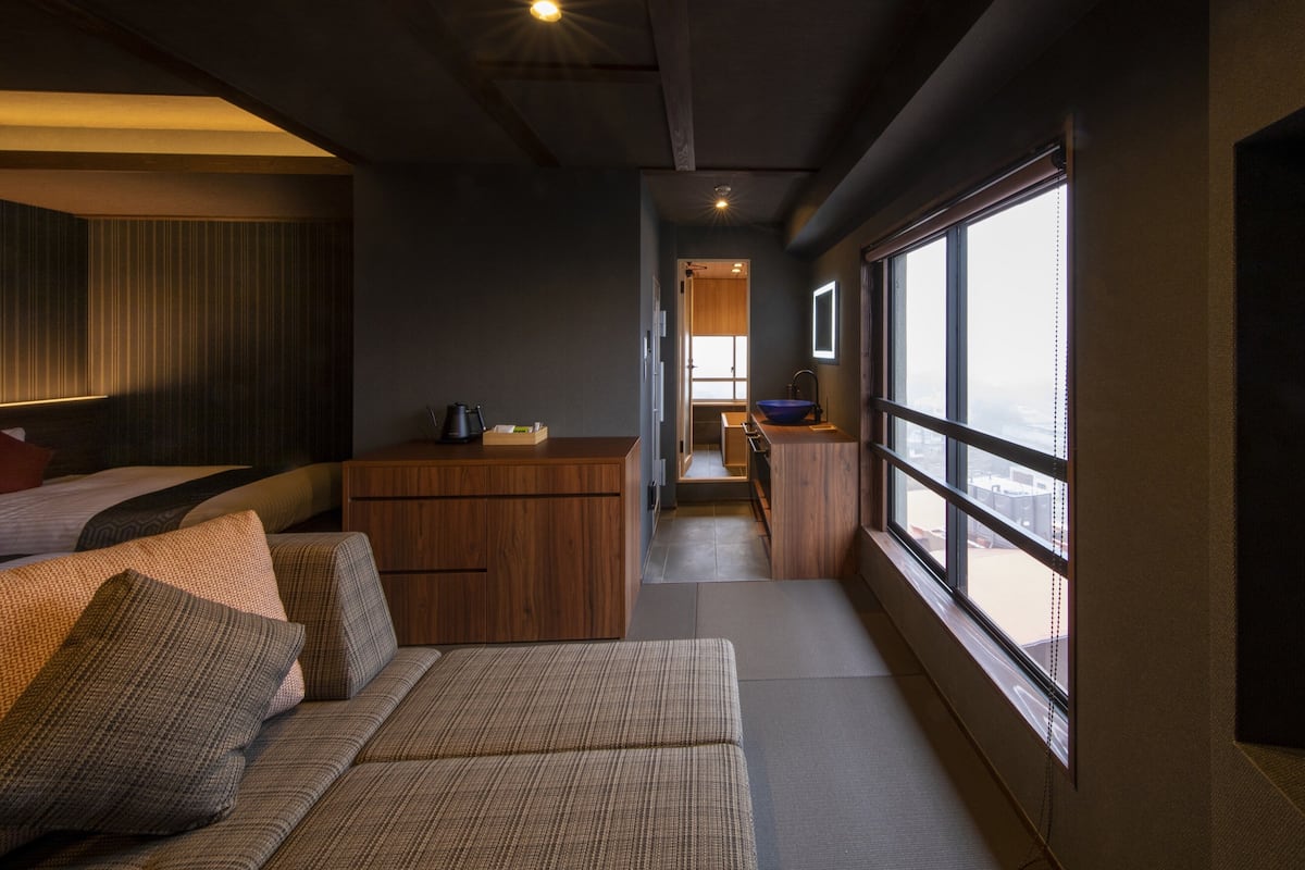 A modern hotel room with a sofa, a wooden cabinet, a small kitchen area, and a large window offering a view.