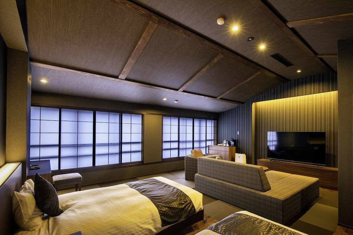 A modern hotel room with a large bed, a sofa, a TV, and a view through large windows.