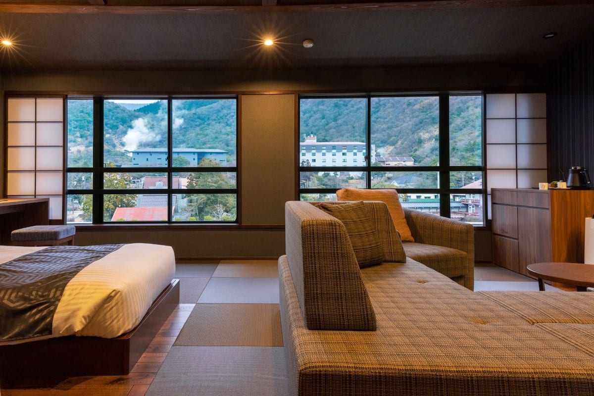 A modern hotel room with a large window, a bed, a sofa, and a small table.