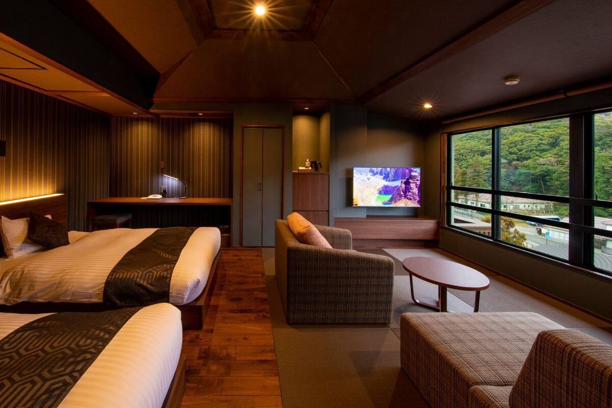 A modern hotel room with two beds, a sofa, a small table, a TV, and a large window offering a view.
