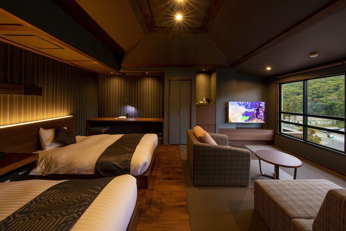 A modern hotel room with two beds, a desk, a TV, and a large window offering a view of the outdoors.