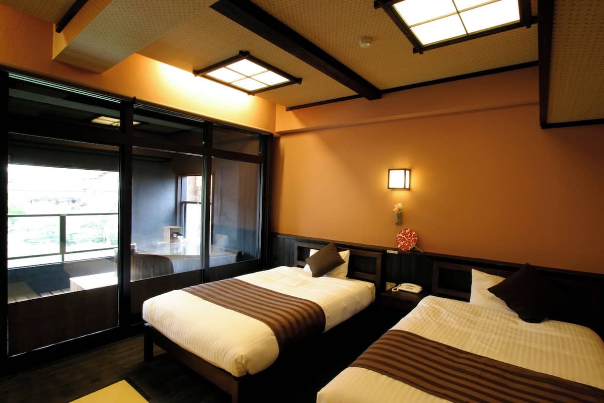 A hotel room with two beds, a large window, a nightstand, and a wall-mounted lamp.