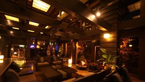 A dimly lit lounge area with wooden furniture, a central fire feature, and a view of the outside through a large window. - Unzen Fukudaya (Unzen)