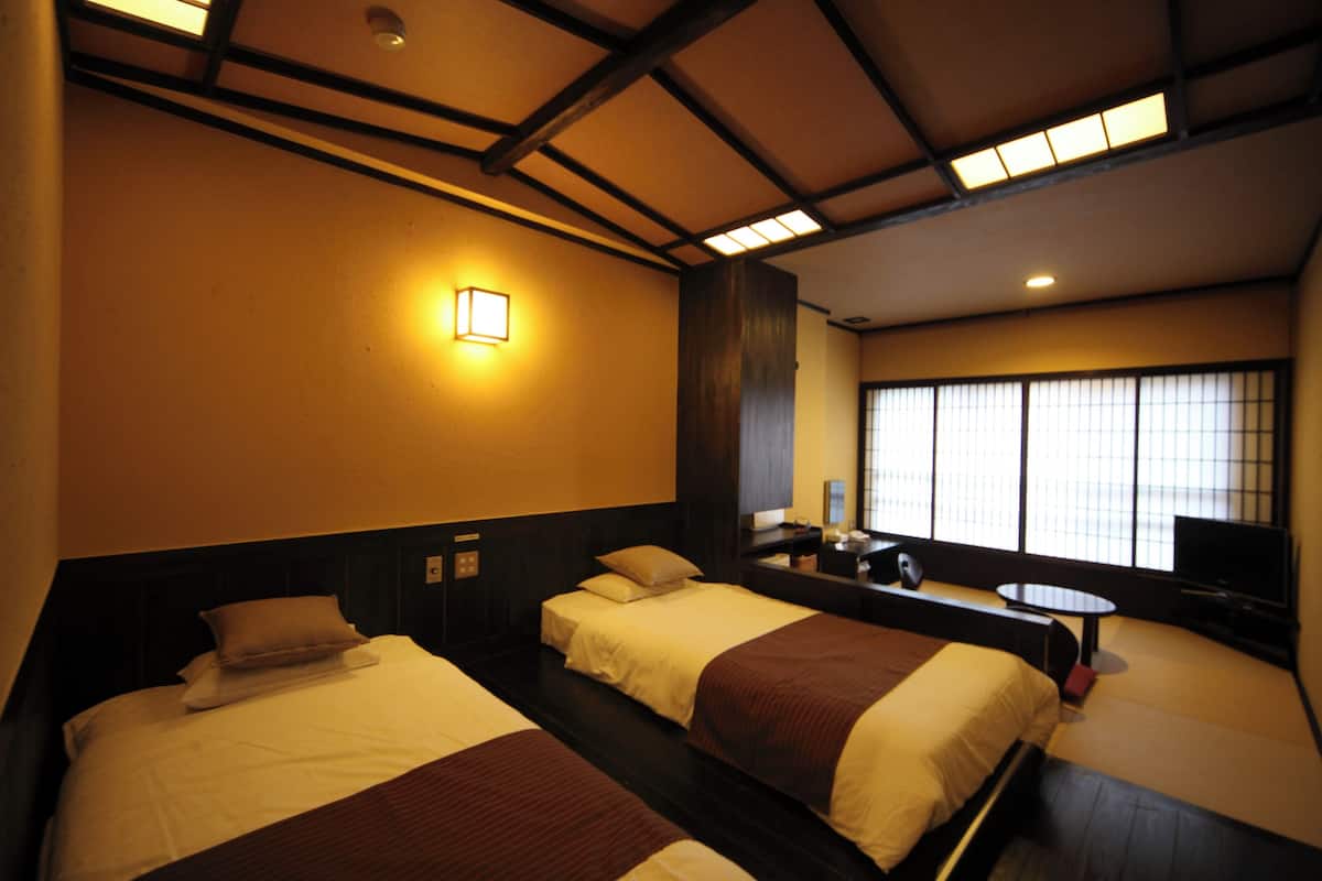 A hotel room with two beds, a small table, a TV, and a window with blinds.
