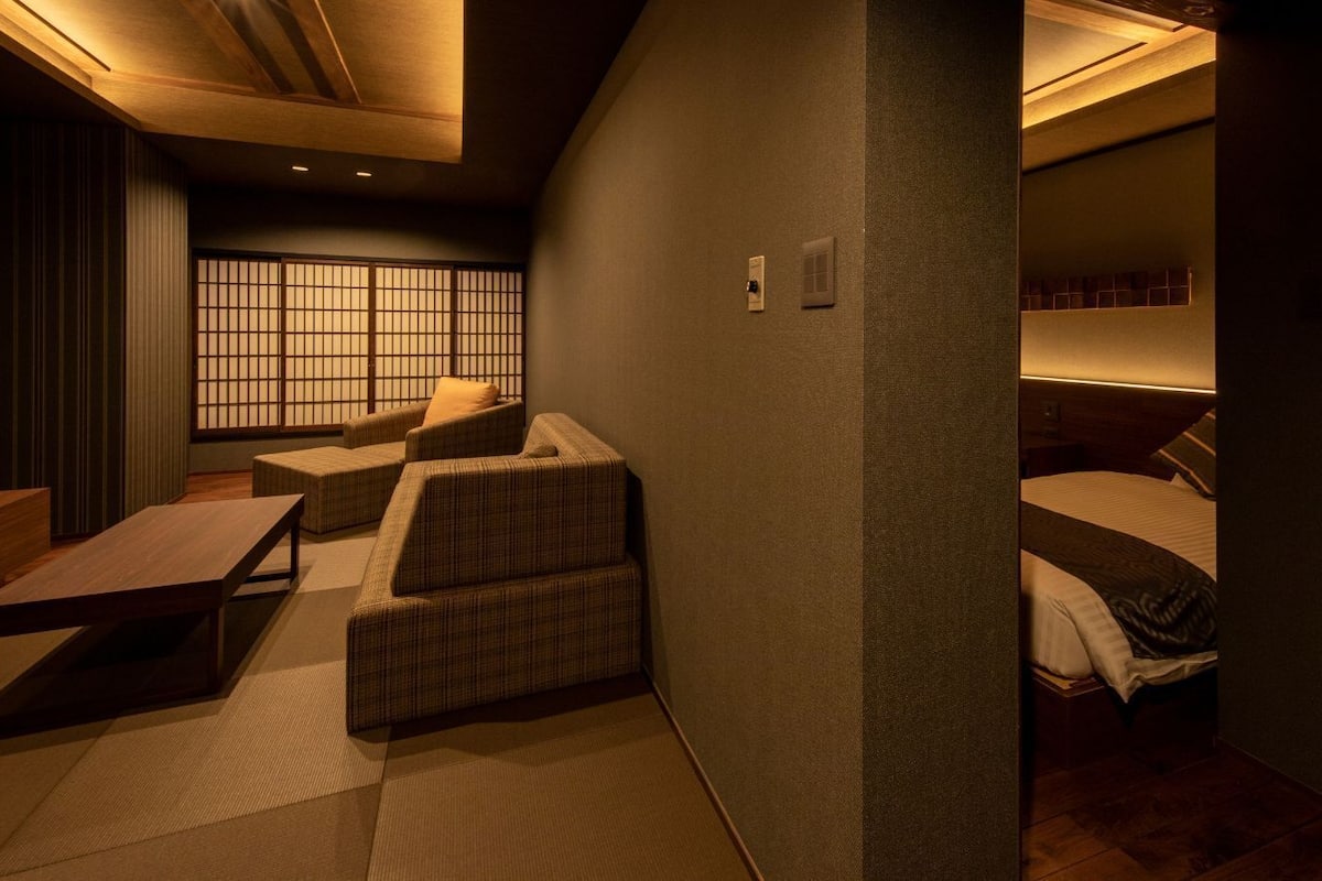 A compact hotel room with a bed, a small sofa, a wooden table, and a tatami mat floor.