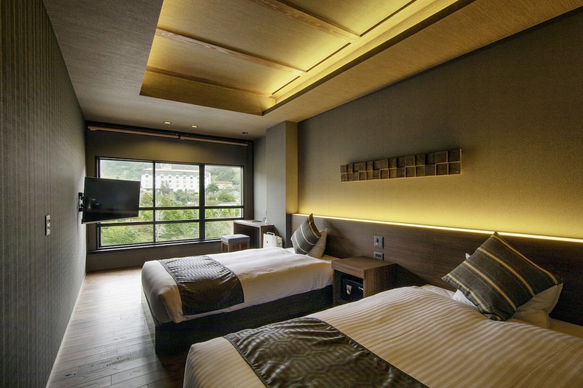 A modern hotel room with two beds, a TV mounted on the wall, a desk, and a window with a view.