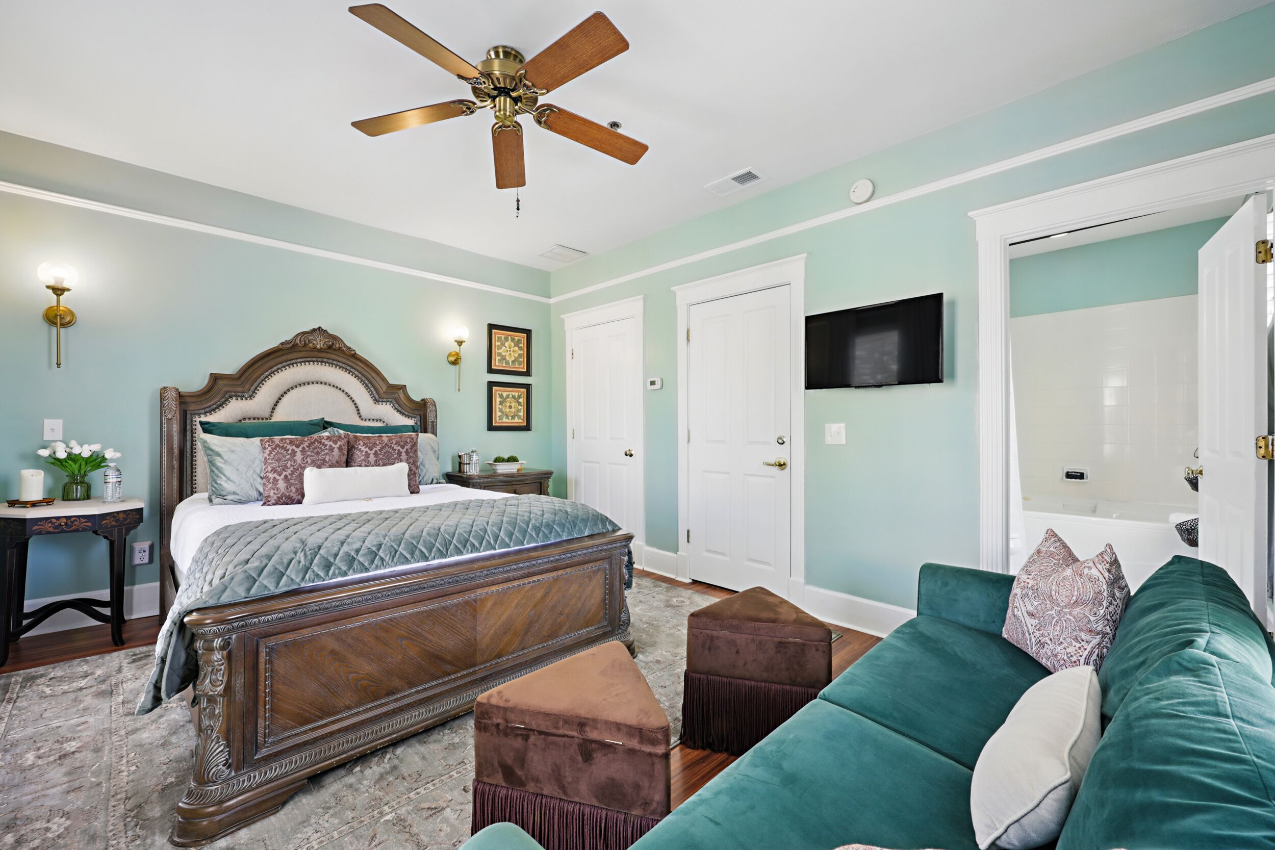 A bedroom with a bed, a green sofa, a ceiling fan, and a television on the wall.