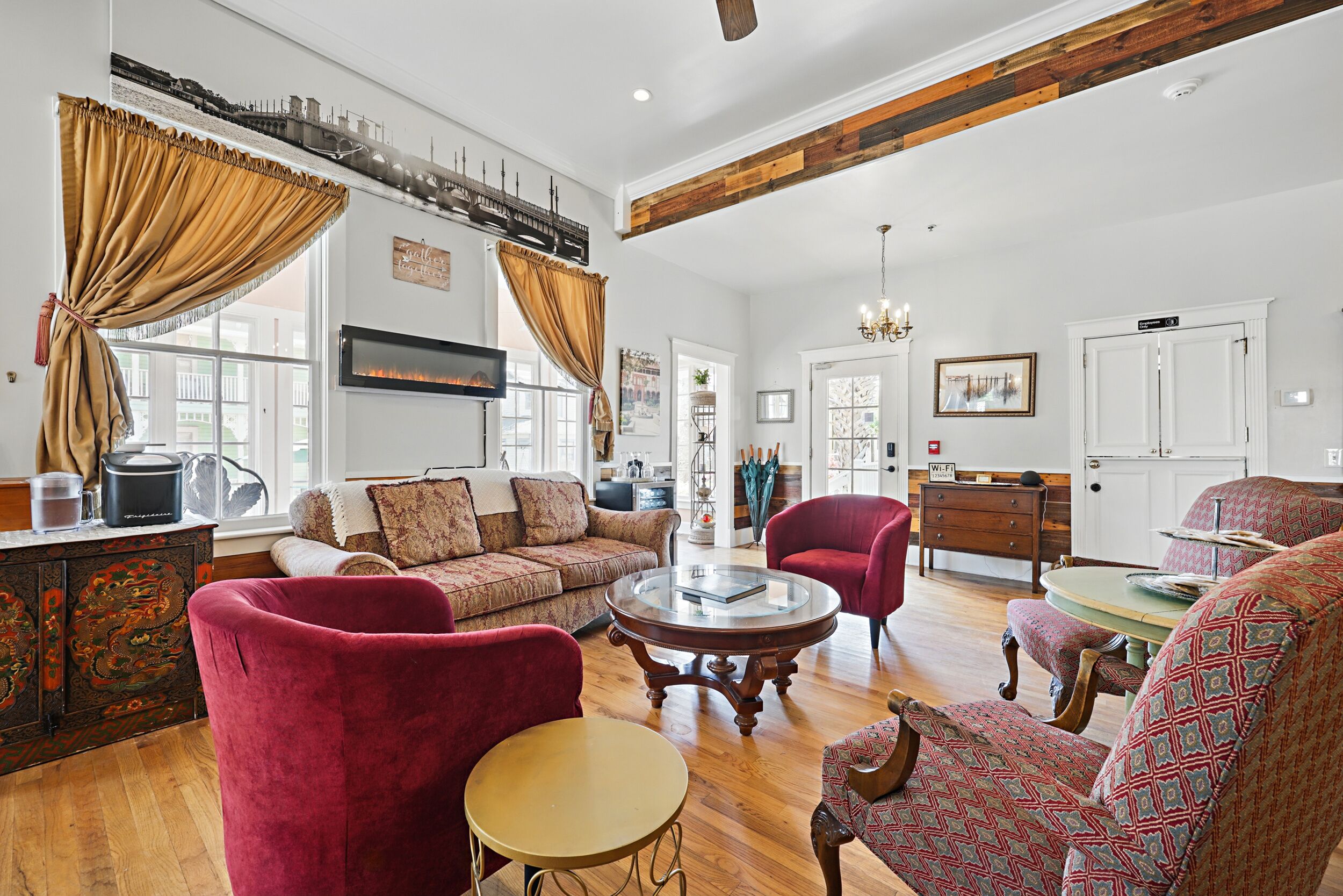 A spacious living room with a fireplace, red velvet chairs, a wooden coffee table, and a large window with curtains.