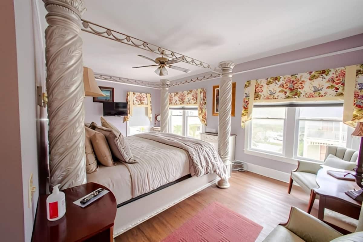 A bedroom with a four-poster bed, a ceiling fan, a desk with a chair, and a window with floral curtains.