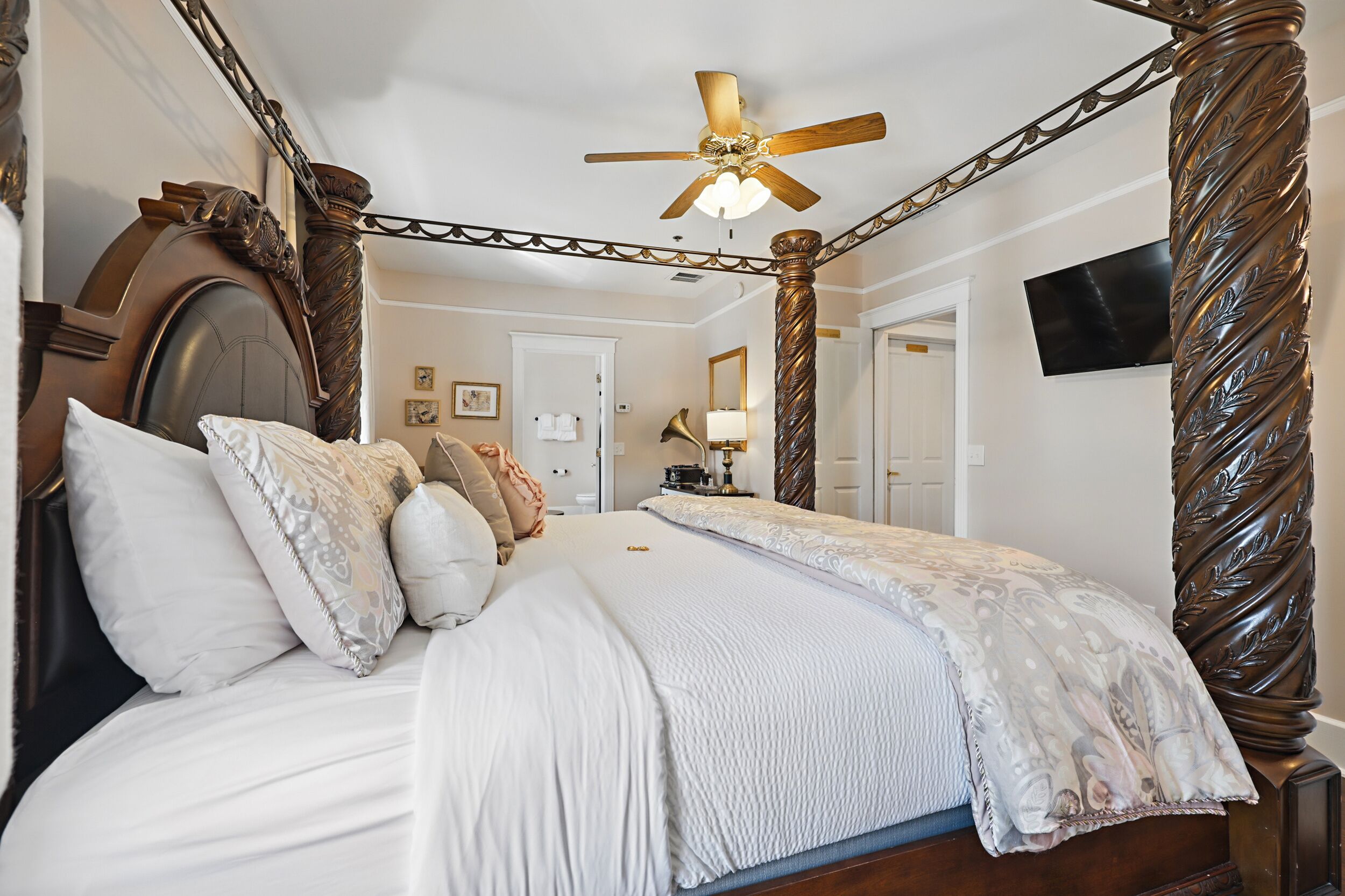 A four-poster bed with a ceiling fan, a television on the wall, and a door leading to another room.