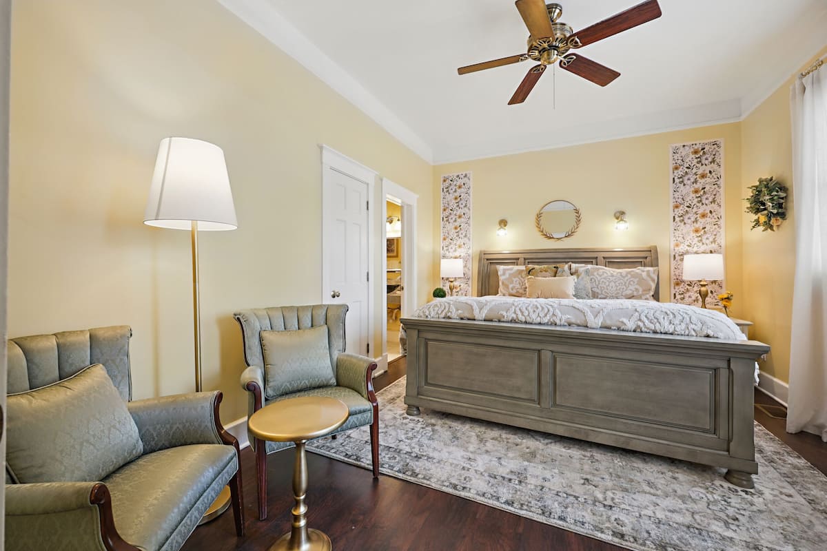 A bedroom with a large bed, a ceiling fan, a chair, a table, and a lamp.