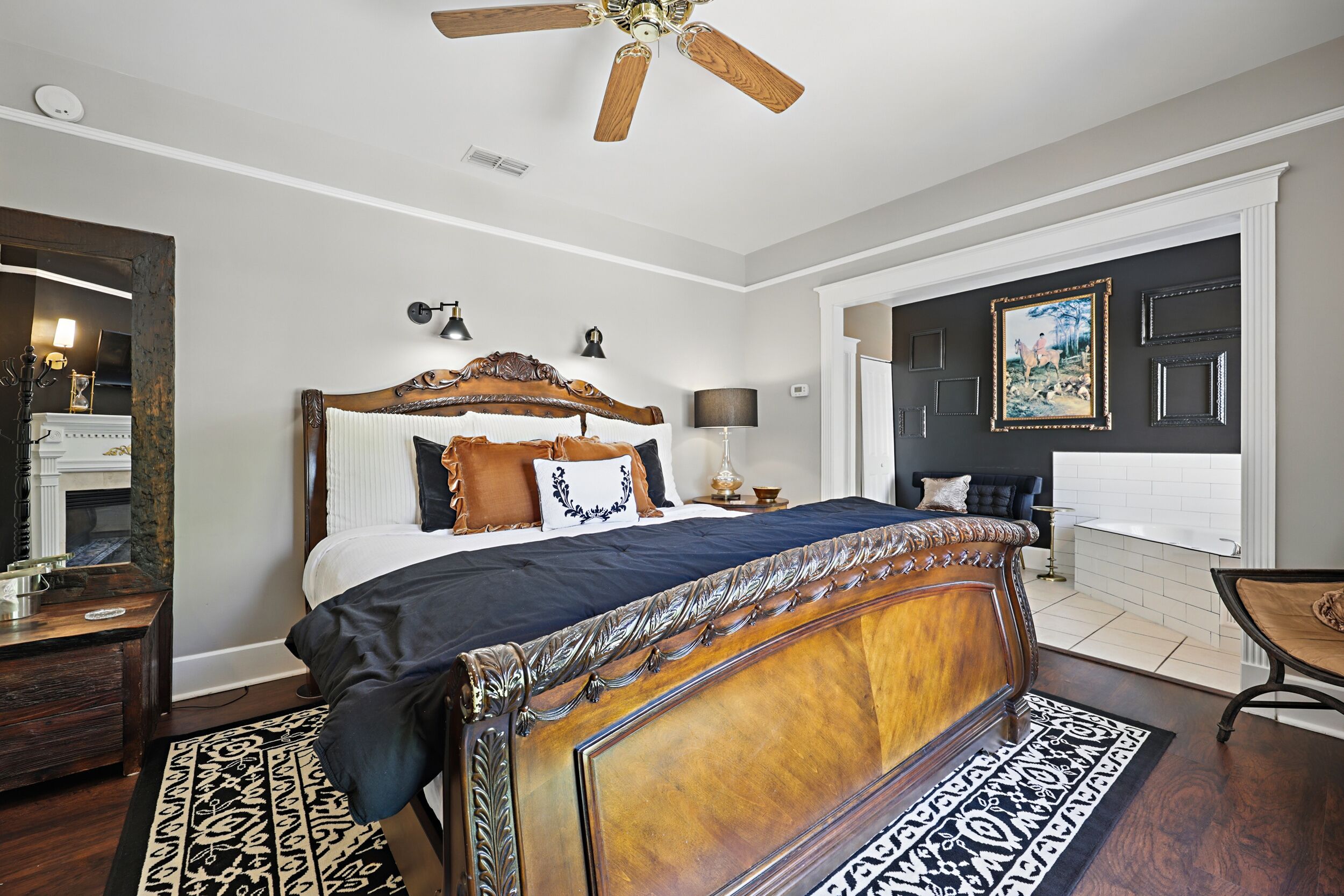 Quarto com cama grande, ventilador de teto, mesa de cabeceira, cômoda, espelho e banheiro com banheira e vaso sanitário visíveis por uma porta aberta.