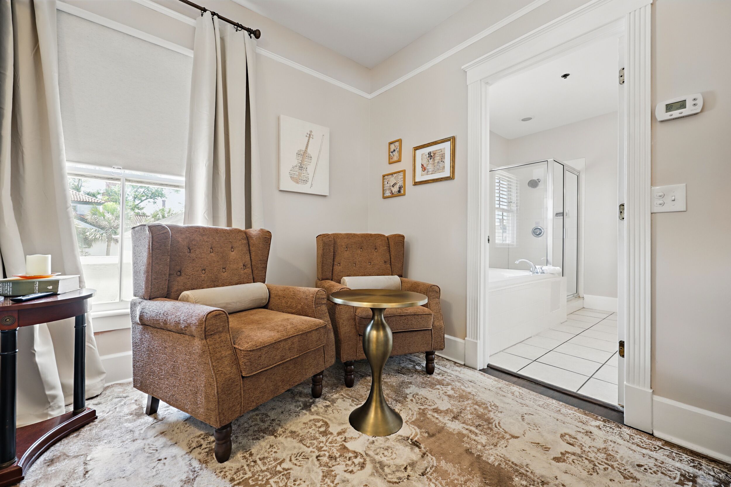 Une pièce avec deux fauteuils marron, une petite table ronde, un tapis à motifs et une vue sur une salle de bain équipée d’une douche et de toilettes.