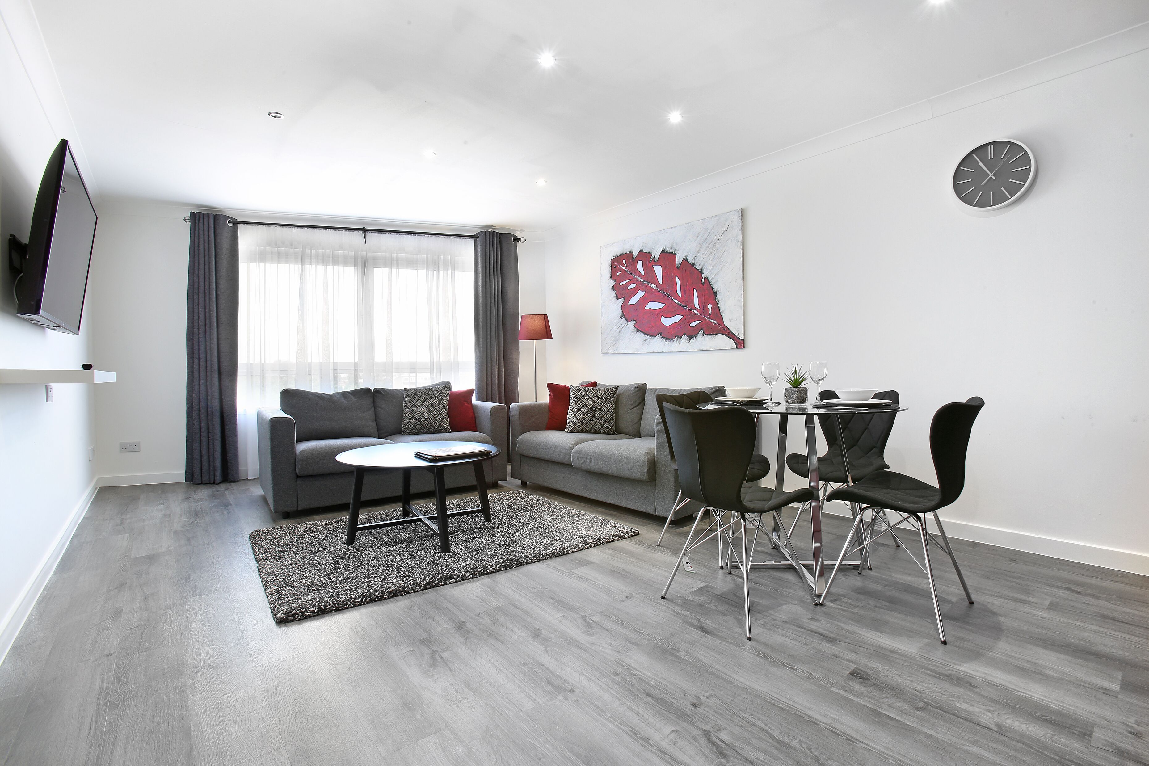 Una sala de estar moderna con un sofá gris, una mesa redonda y un comedor con mesa de vidrio y sillas metálicas.
