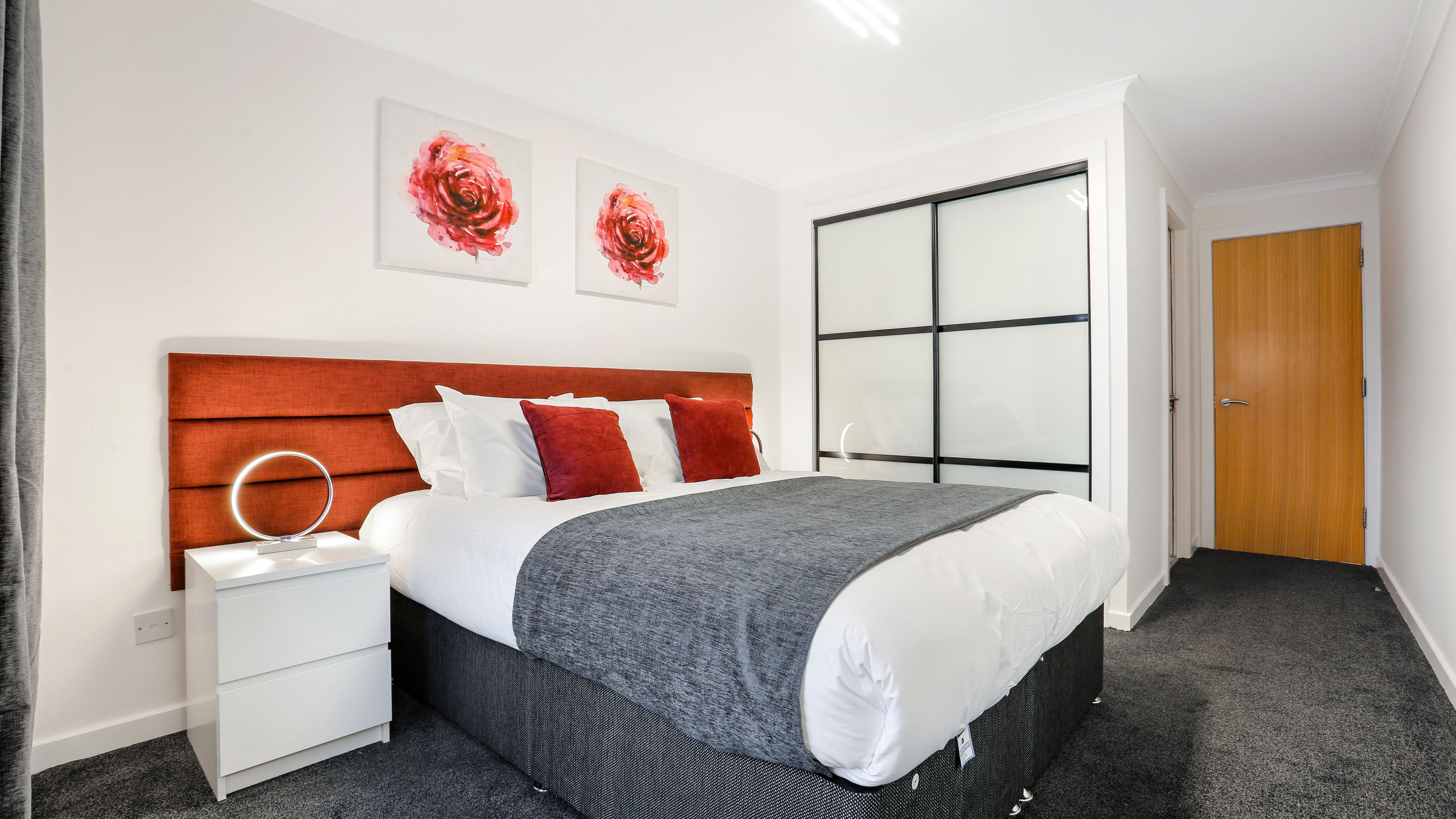 A modern hotel room with a large bed, a bedside table with a lamp, and a large mirror. There are two red floral paintings on the wall and a sliding glass door to the right.