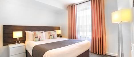 A hotel room with a large bed, a nightstand with a lamp, a floor lamp, and a window with curtains.