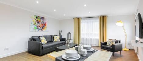 A modern living room with a black sofa, a dining table set with white plates and cups, a yellow floor lamp, and a colorful abstract painting on the wall.