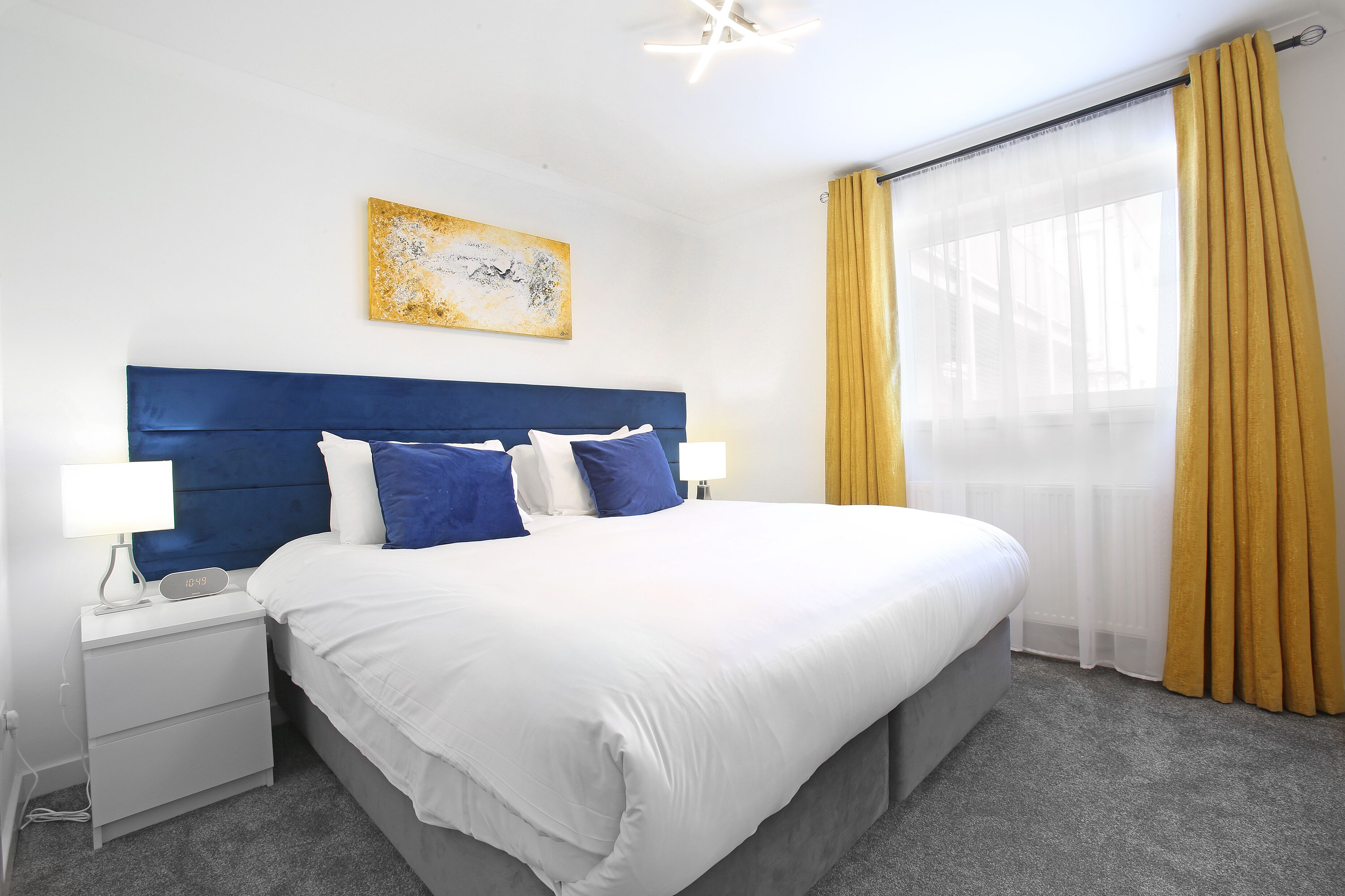 A hotel room with a large bed, a nightstand with a lamp, a wall clock, and a window with yellow curtains.
