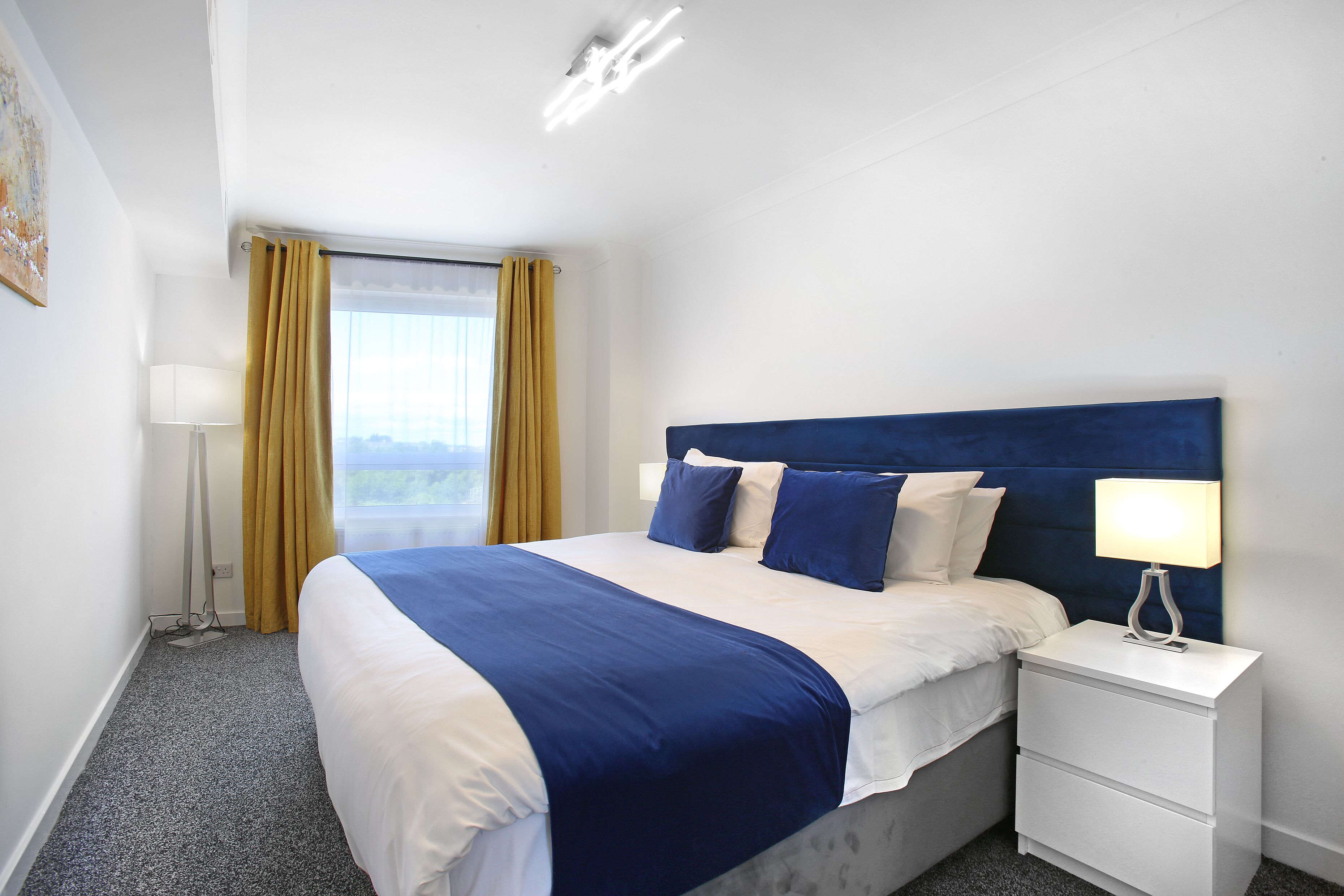 Quarto de hotel com cama, mesa de cabeceira, abajur e janela com cortinas.
