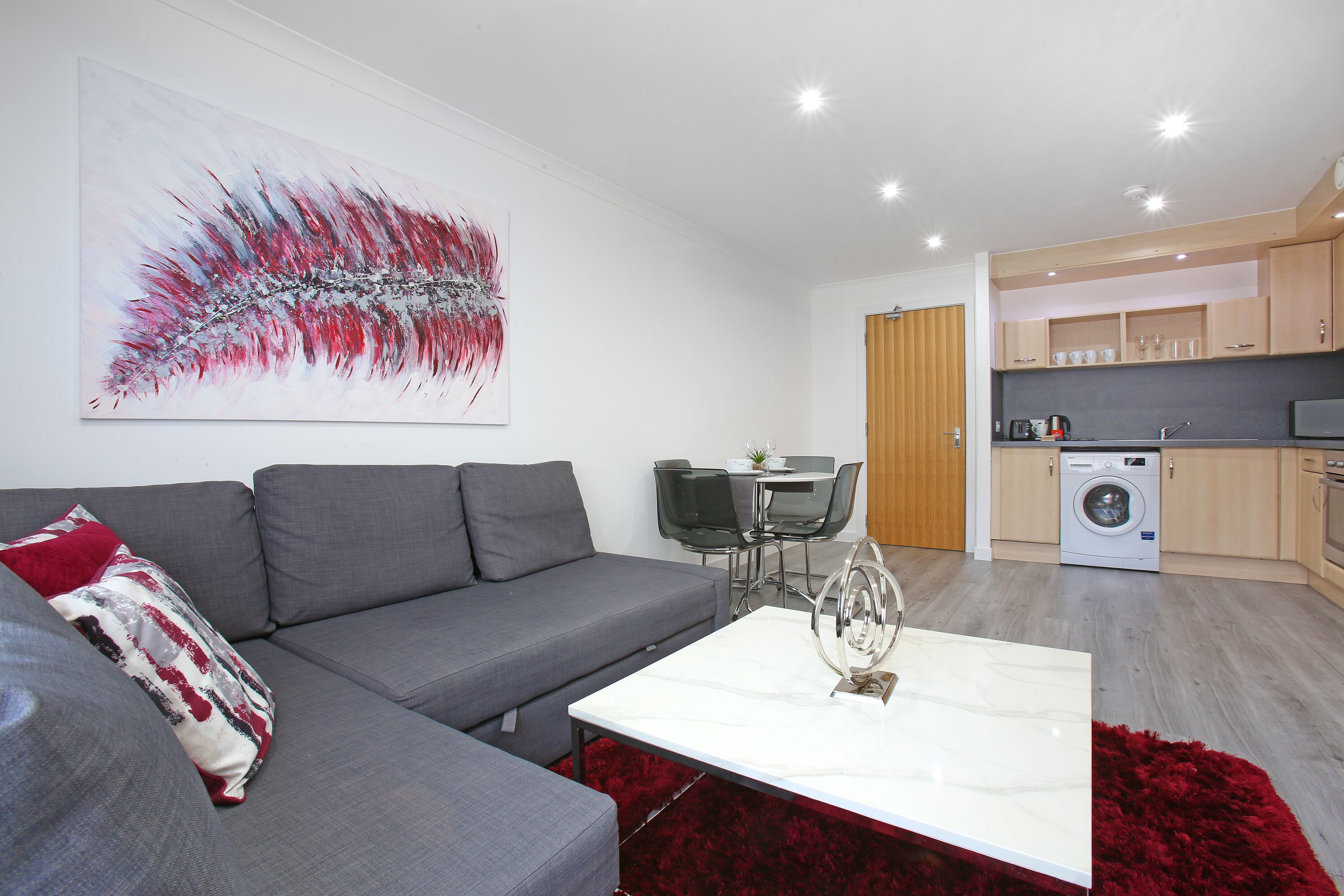 Ein modernes Wohnzimmer mit einem grauen Sofa, einem weißen Couchtisch und einem roten Teppich. Es gibt eine Essecke mit Tisch und Stühlen sowie eine Küche mit Holzschränken und einer Waschmaschine.