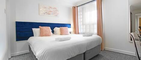 A hotel room with a large bed, a blue headboard, and a window with curtains.