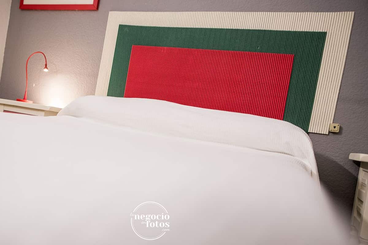A neatly made bed with white linens, a red lamp on a bedside table, and a geometric-patterned wall hanging.