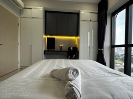 A hotel room with a large bed, a dark headboard, a small desk with a chair, a mirror, and a view of a cityscape through a large window.