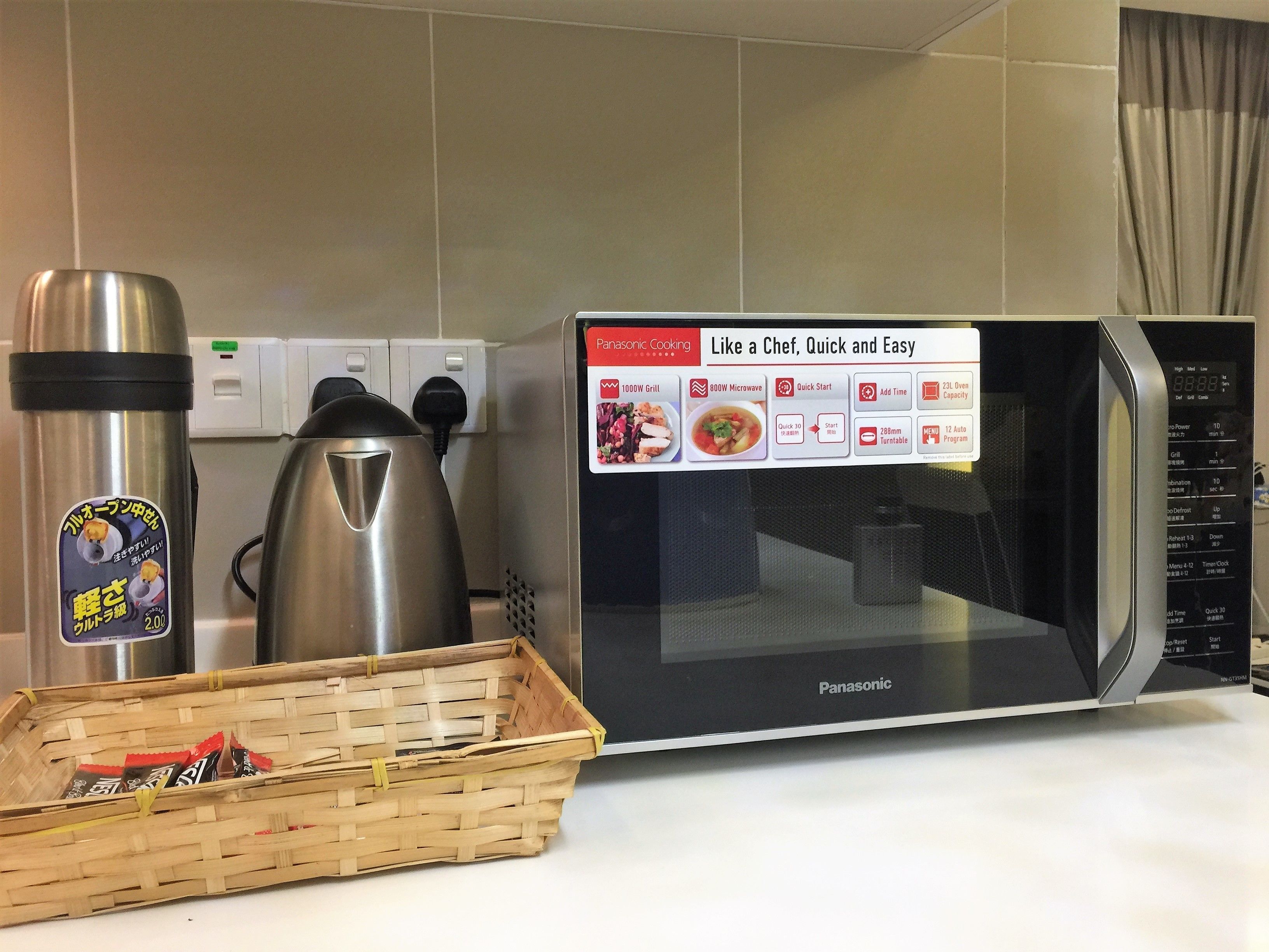 Una cocina con un microondas Panasonic, un hervidor de acero inoxidable y una cesta con alimentos.