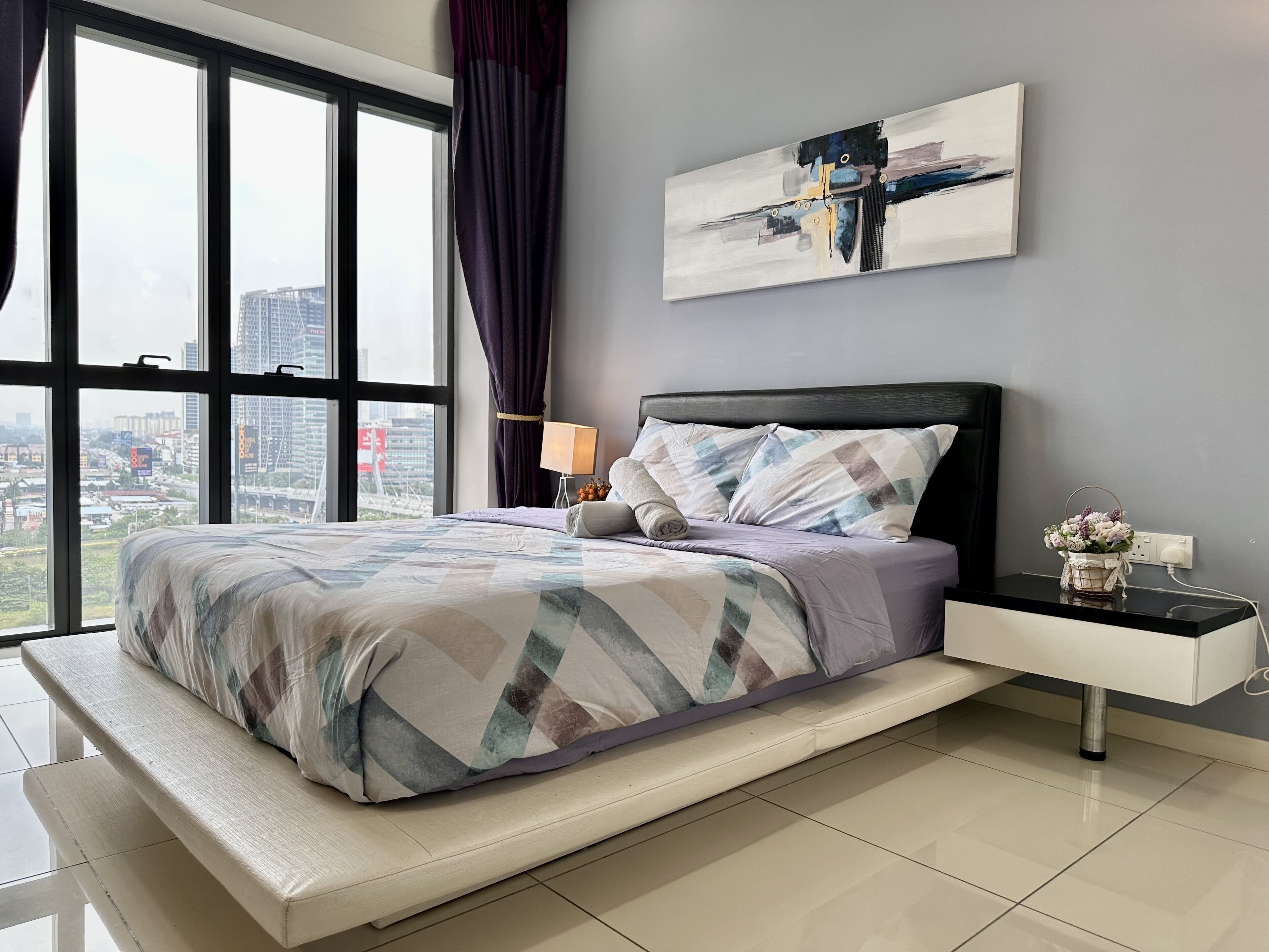 A modern bedroom with a large bed, a nightstand, a lamp, and a city view through large windows.