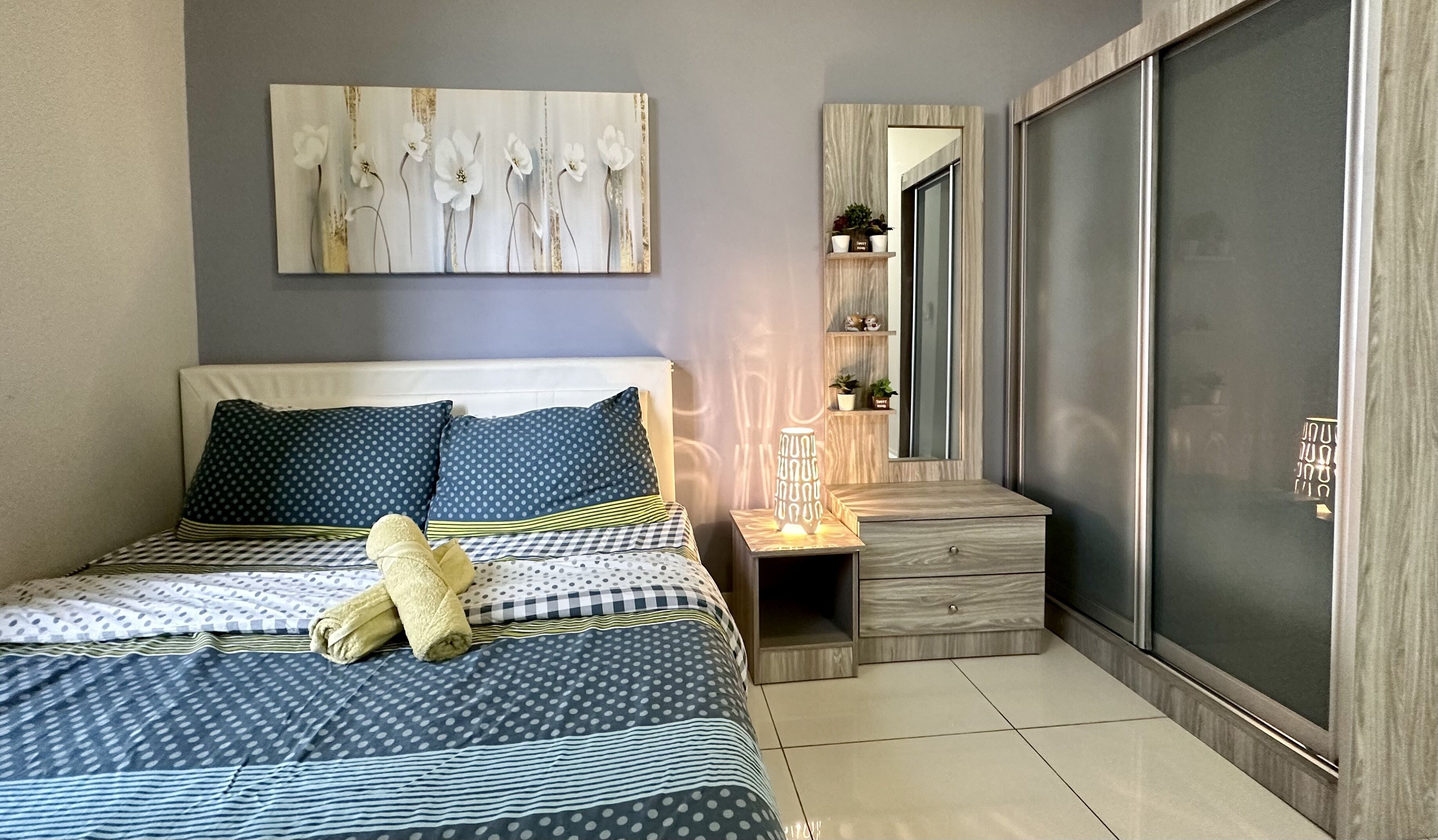 Un dormitorio moderno con una cama, almohadas, una mesita de noche con lámpara, un espejo y un armario.