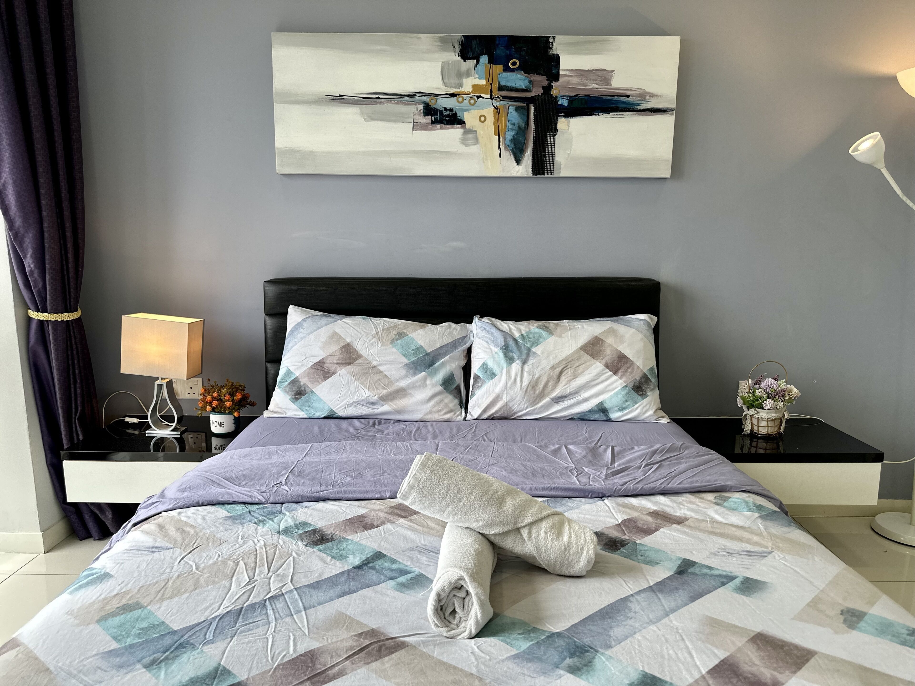 A neatly made bed with a patterned bedspread, a pillow, and a rolled towel. There is a bedside table with a lamp and a small decorative item. A painting is hung above the bed.