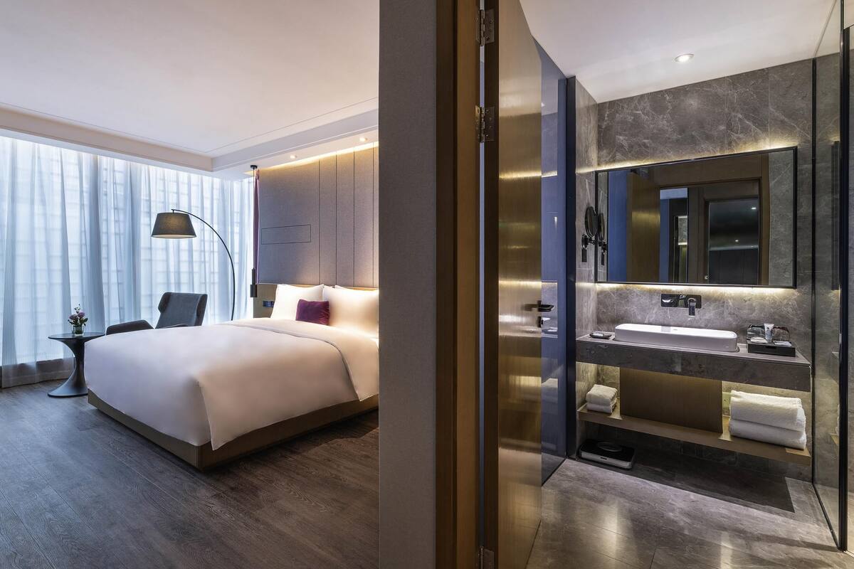 A modern hotel room with a large bed, a separate bathroom with a large mirror, and a view of the city through the window.