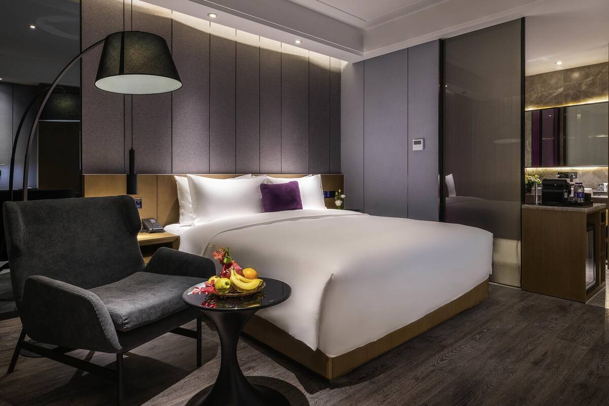 A modern hotel room with a large bed, a desk with a lamp, a chair, and a small table with fruit.