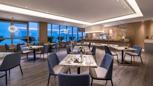 A modern dining area with multiple tables and chairs, large windows with a view, and a kitchen area in the background. - Mercure Kunshan MIXC Hi-tech (Suzhou)