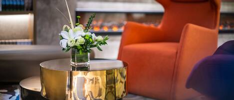 Un salón moderno con una mesa redonda dorada, un jarrón blanco con flores, un sillón naranja y un televisor al fondo.