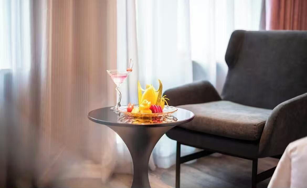 A modern room with a dark grey sofa, a small round table with a martini glass and fruit, and a window with curtains.