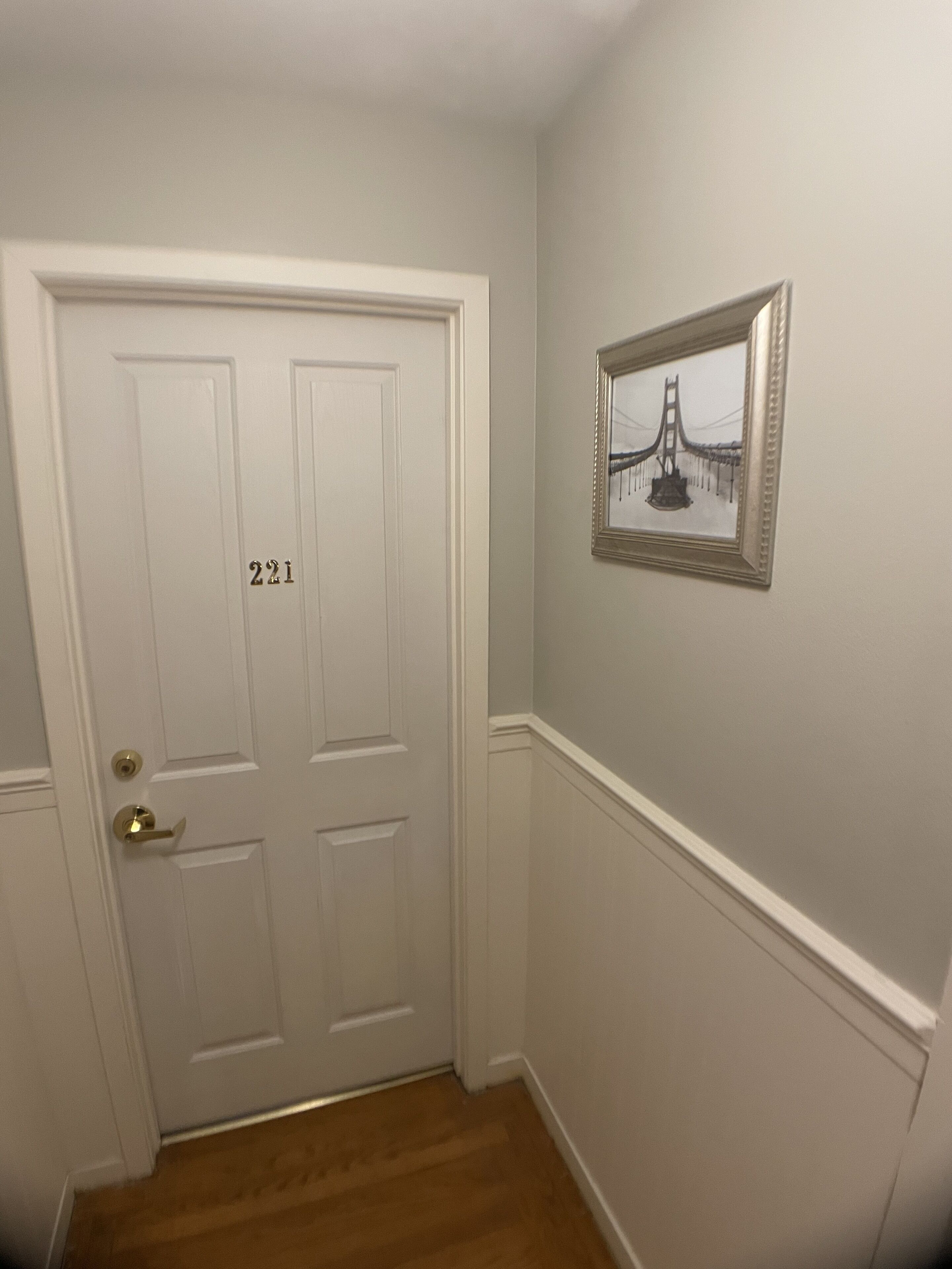 Een gang met een witte deur waarop “221” staat, een ingelijst foto van een brug aan de muur, en houten vloeren.