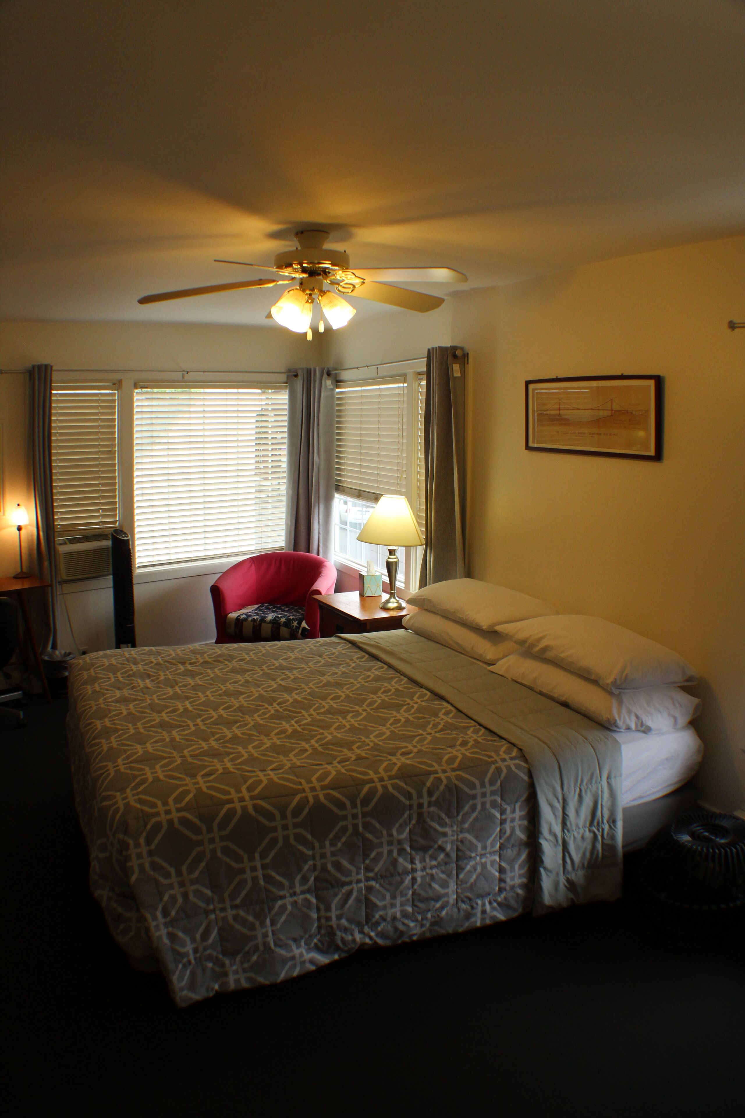 A hotel room with a bed, a red chair, a desk with a lamp, and a ceiling fan.