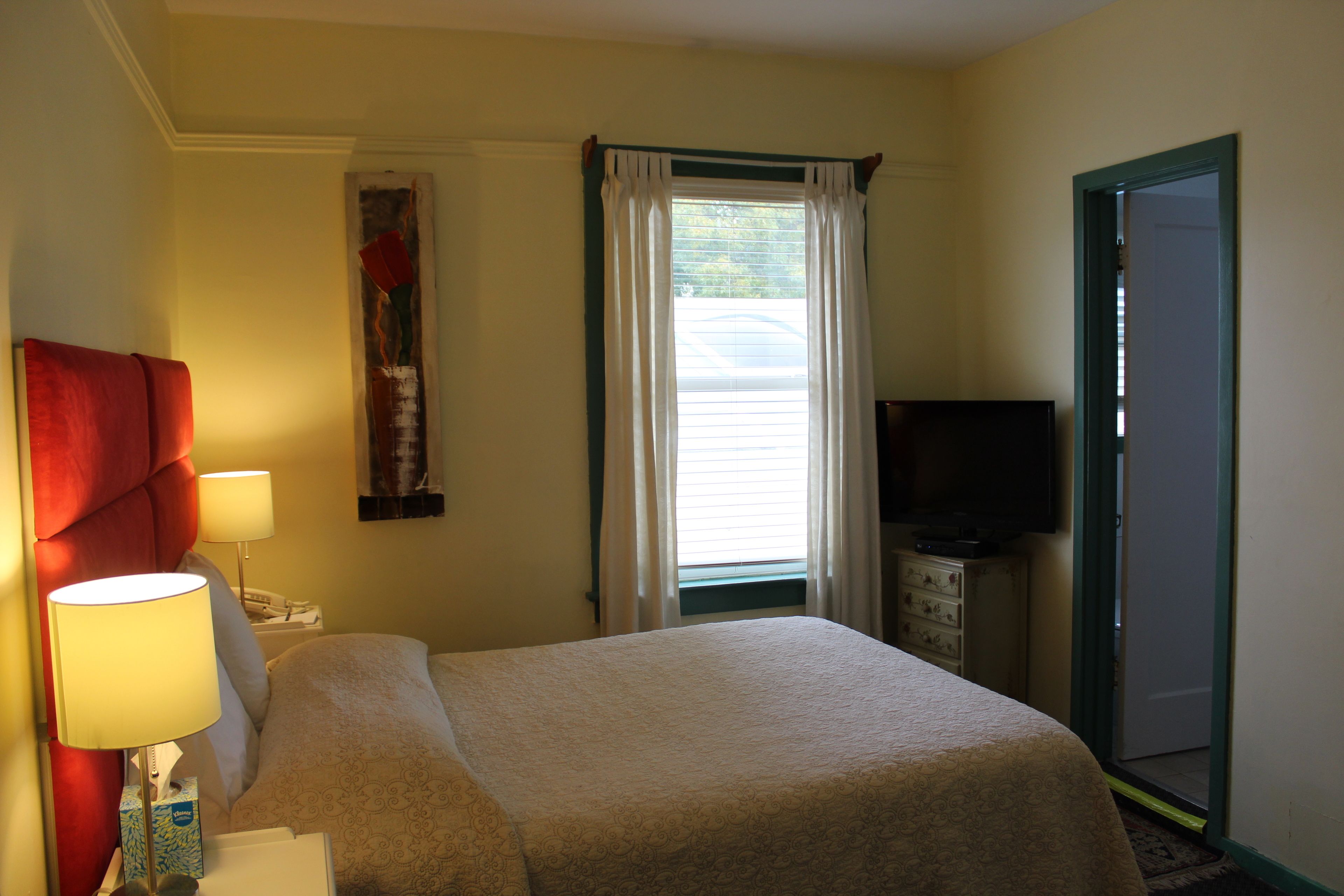 Una habitación de hotel con una cama, televisión, mesita de noche, lámpara y ventana con cortinas.