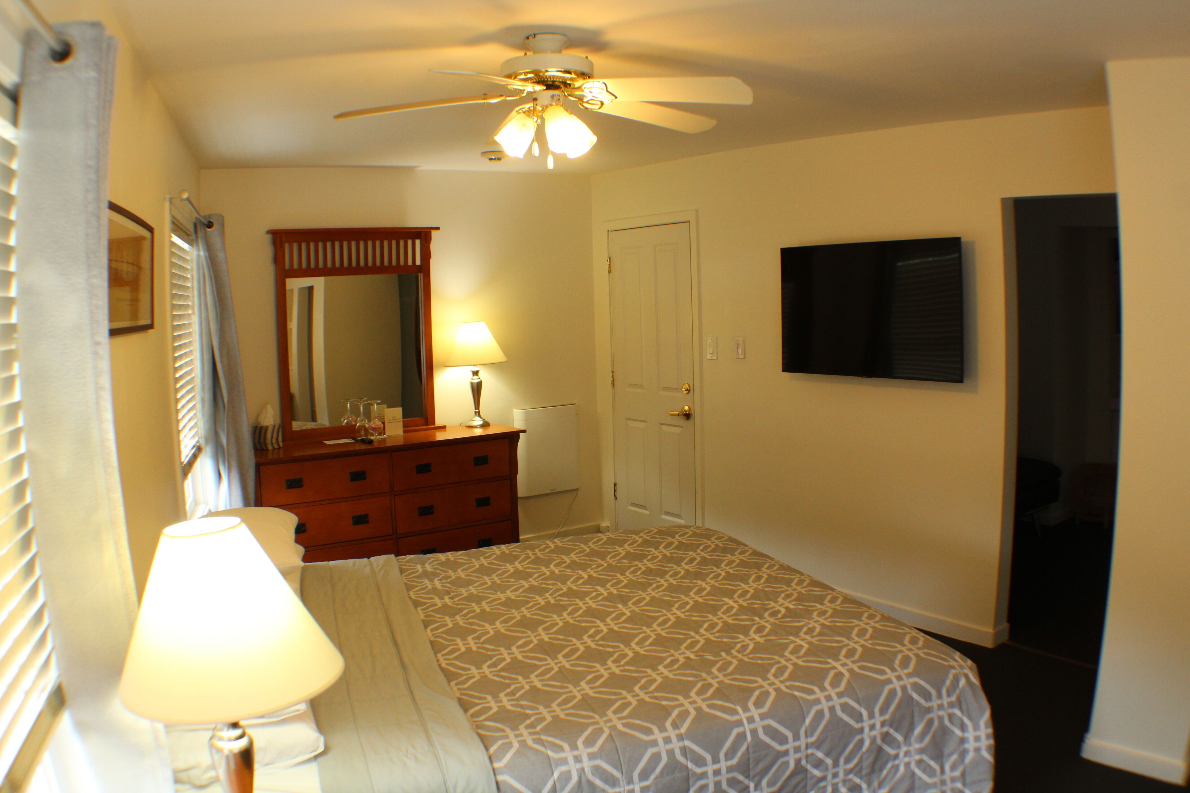A bedroom with a bed, a dresser, a mirror, a ceiling fan, and a TV on the wall.