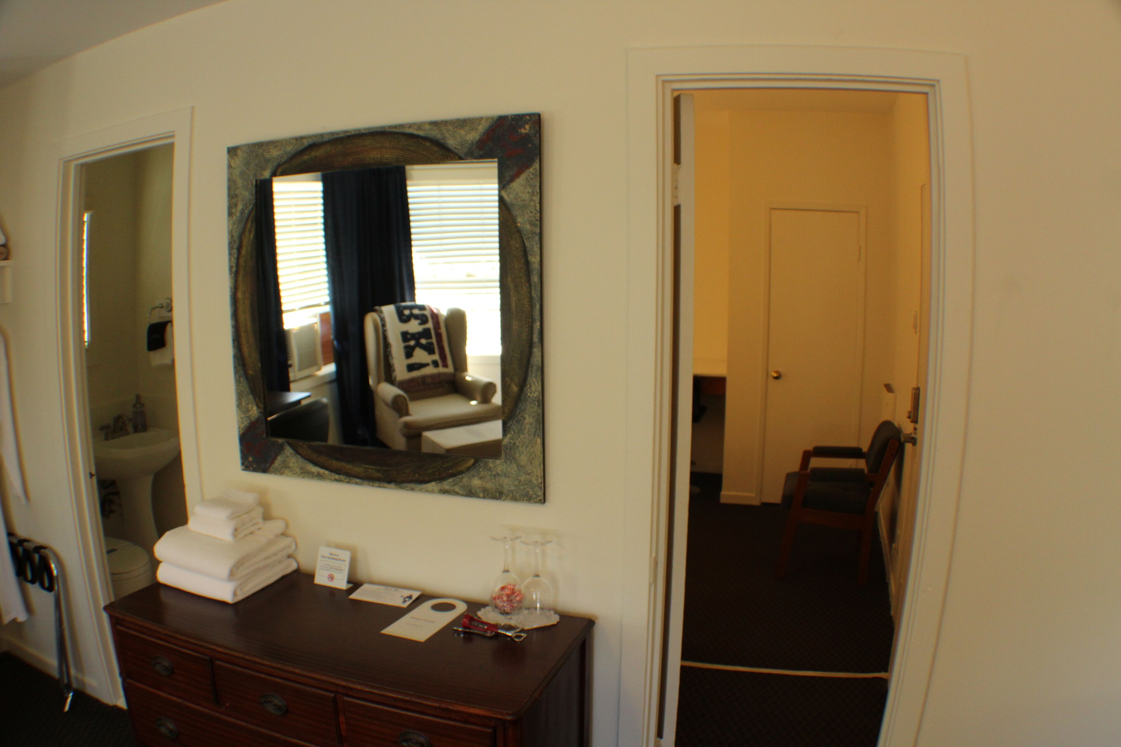 A hotel room with a wooden dresser, a mirror, a chair, and a door leading to another room.