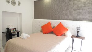 Ein Hotelzimmer mit einem großen Bett, zwei orangefarbenen Kissen, einem kleinen Tisch mit Lampe und einer Wand mit Dekorationselementen.