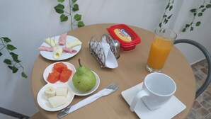 Ein Tisch mit Frühstücksangeboten, darunter ein Birne, Wassermelone, Bananen und Schinken. Ein Glas Orangensaft und eine weiße Tasse sind ebenfalls vorhanden.