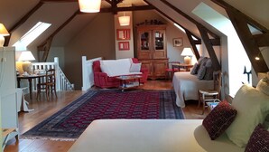 A cozy loft bedroom with a high ceiling, exposed wooden beams, a large bed, a red armchair, a wooden cabinet, and a dining area with a table and chairs. - La Peuterne (Mouazé)