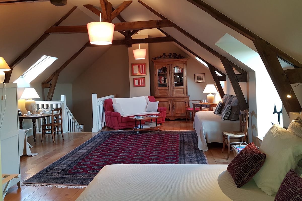 A cozy loft bedroom with a high ceiling, exposed wooden beams, a large bed, a red armchair, a wooden cabinet, and a dining area with a table and chairs.