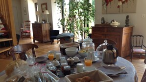 A breakfast table set with various food items and beverages, surrounded by wooden furniture and a large plant. - La Peuterne (Mouazé)