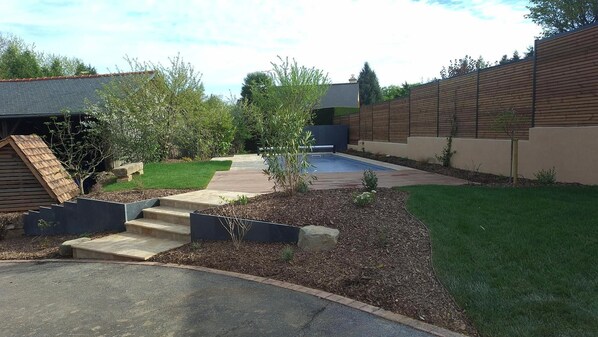 Un patio trasero con piscina, terraza de madera y un pequeño jardín.
