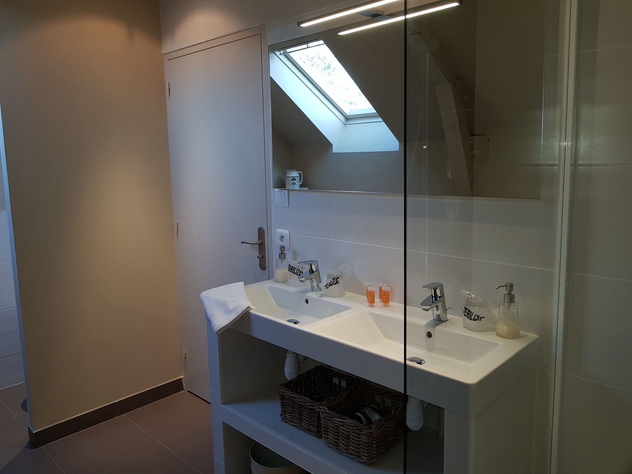 A modern bathroom with a skylight, a white sink with two faucets, and a glass shower enclosure.