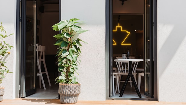 Ein moderner Eingangsbereich mit Blumentopf, einer Neonreklame und einem Essbereich, der durch die Glastüren sichtbar ist.