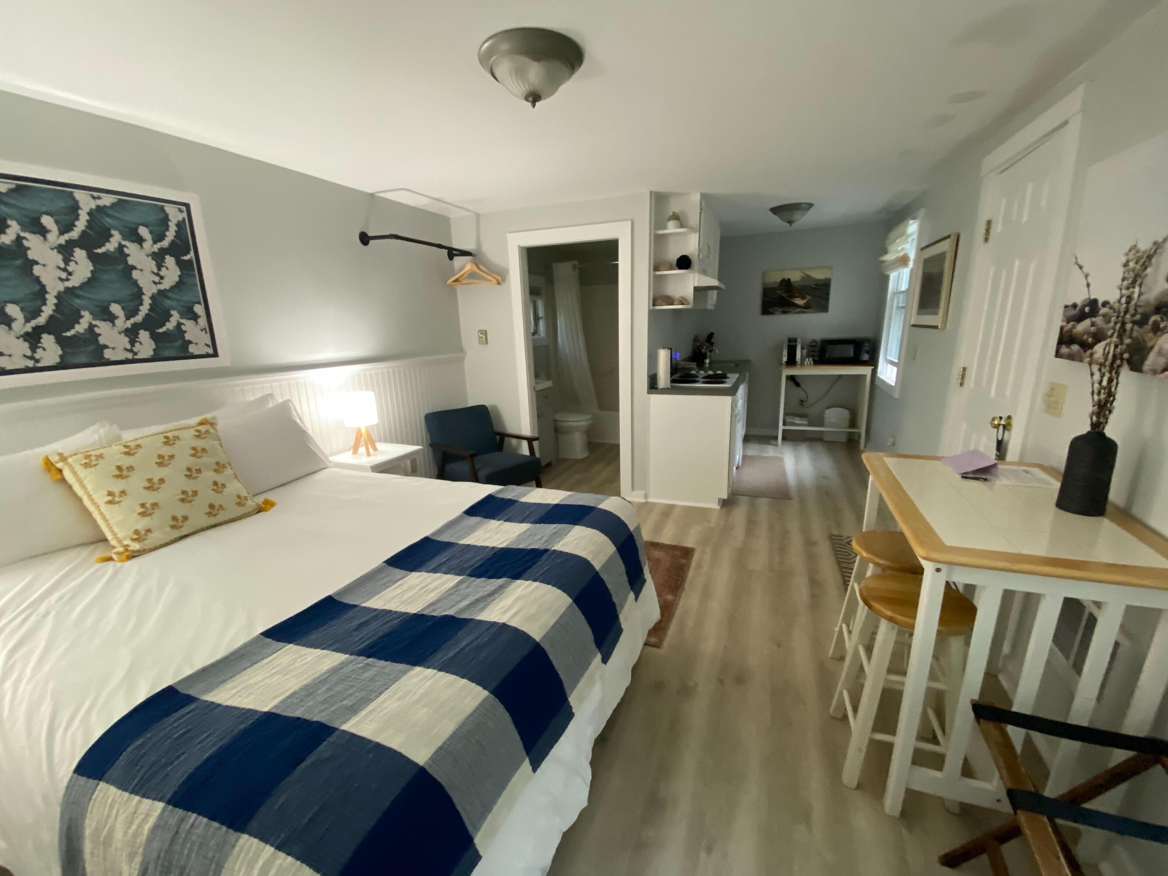 A hotel room with a bed, a desk, a chair, a kitchenette, and a bathroom.